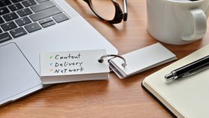 There's a laptop computer, a notebook, a pen and an open vocabulary book on the desk. The word Content Delivery Network is written on it.