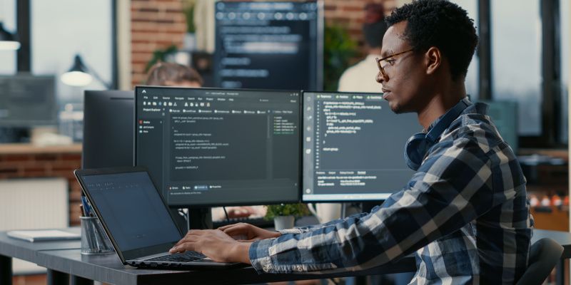 A male sysadmin working on a laptop with two monitors in the background.